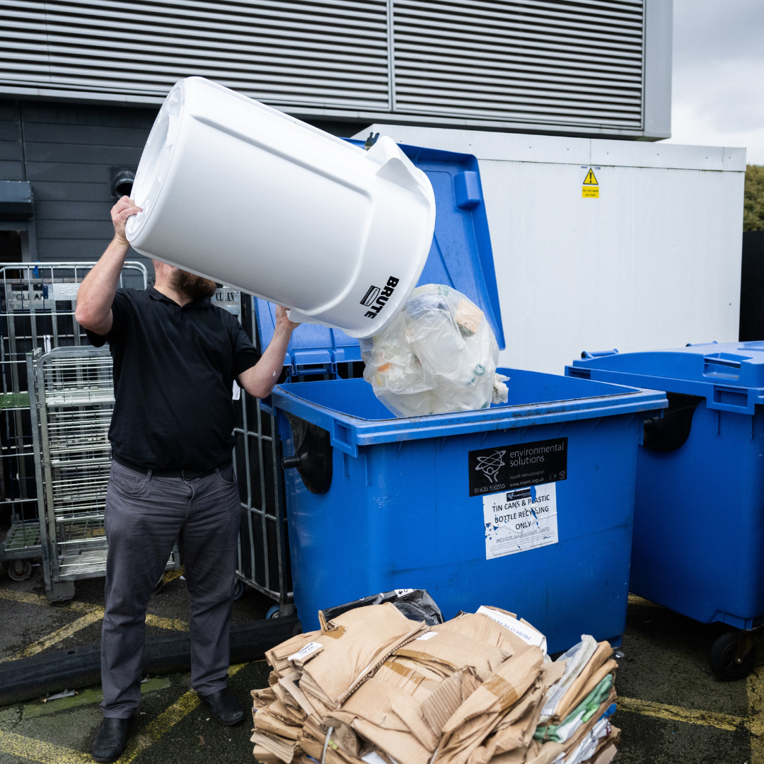 Person emptying white bin into large blue recycling dumpster outdoors.